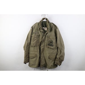 May include: An olive green field jacket featuring a high collar, a front zipper closure, and four large button-flap pockets. The chest displays a name patch reading MAIER and a circular patch with the text SEABEES and a bee graphic illustration.