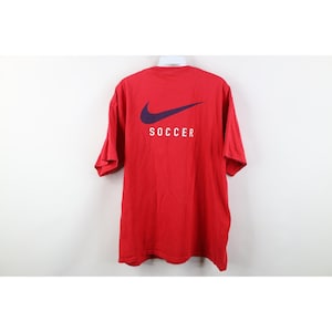 May include: A red t-shirt with a purple Nike swoosh logo and the word "SOCCER" in white. The shirt is short-sleeved and appears to be made of cotton. The shirt is hanging on a white hanger.