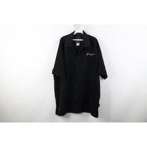 May include: Black short-sleeve button-up shirt with a collar and two chest pockets. The shirt has a small white embroidered signature on the right chest. The sleeves are rolled up. The shirt is hanging on a white hanger.