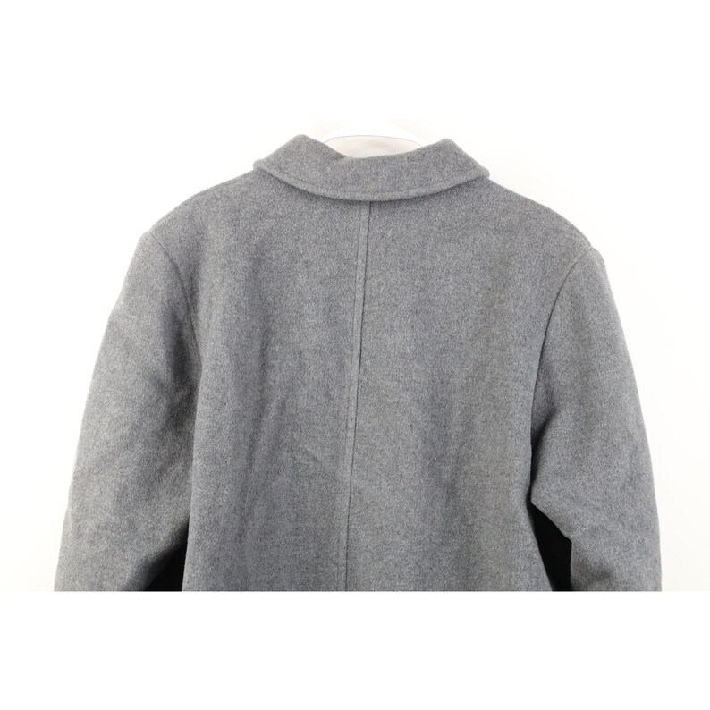 May include: A gray wool coat, viewed from the back, with a classic collar and a center seam. The coat is hanging on a white hanger. The fabric has a subtle texture.