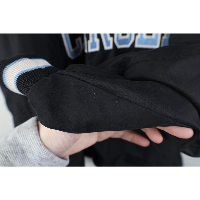 May include: Black jacket with the word "UNC" in light blue and white lettering. The jacket has a ribbed cuff with blue and white stripes. The sleeve is black.