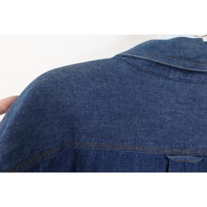 May include: Close-up of a blue denim jacket. The jacket features a collar, shoulder seams, and a pocket detail. The stitching is a contrasting brown color, and the fabric has a textured appearance.