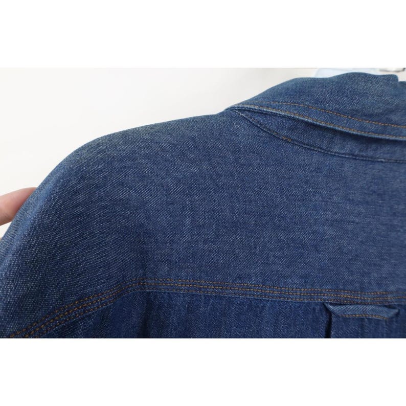 May include: Close-up of a blue denim jacket. The jacket features a collar, shoulder seams, and a pocket detail. The stitching is a contrasting brown color, and the fabric has a textured appearance.