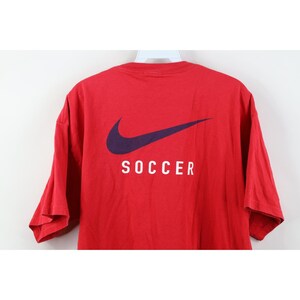 May include: Red short-sleeve t-shirt with a large purple Nike swoosh logo and the word "SOCCER" in white block letters. The shirt is hanging on a white hanger.