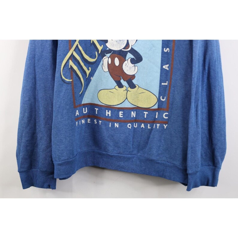 Op de afbeelding: Een blauw sweatshirt met lange mouwen en een afbeelding van Mickey Mouse in een rechthoekig kader. De tekst luidt Mickey, Classic, Authentic en Finest in Quality. Het ontwerp heeft een lichtblauwe achtergrond, bruine randen en witte letters.