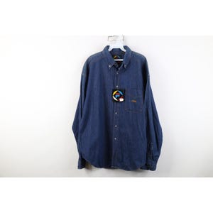 May include: A dark blue denim button-down shirt with long sleeves. The shirt has a button-down collar, a chest pocket, and a small embroidered logo. A tag with a logo is attached to the shirt.