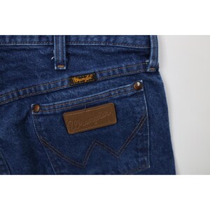 May include: Close-up of dark blue denim jeans with a brown leather patch and a small tag, both displaying the "Wrangler" logo. The jeans have a classic five-pocket design with copper-colored rivets and stitching.