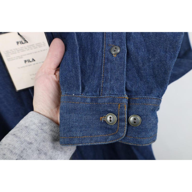 May include: Close-up of a blue denim shirt cuff with two buttons. The cuff is stitched with a contrasting orange thread. A tag with the brand name "FILA" is visible.