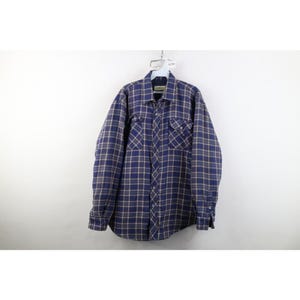 May include: A long-sleeve button-up shirt featuring a plaid pattern in shades of navy blue, tan, and white. The shirt includes a standard collar, a full-length front button closure, and two chest pockets with buttoned flaps on the front panels.
