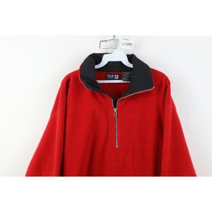May include: A red FILA fleece pullover with a black collar and a silver zipper. The FILA logo is visible on the collar. The pullover is hanging on a white hanger.