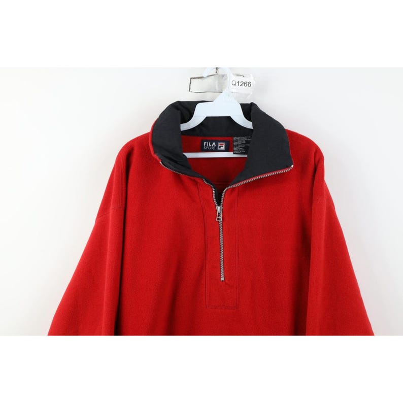 May include: A red FILA fleece pullover with a black collar and a silver zipper. The FILA logo is visible on the collar. The pullover is hanging on a white hanger.