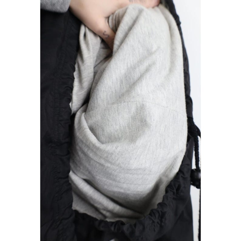 May include: Close-up of a black jacket with a gray interior lining. The jacket has a drawstring closure and a hood. The lining is a soft, light gray fabric. The jacket is made of a smooth, black material.