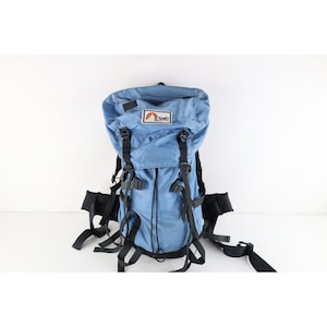 May include: A light blue backpack featuring a black base and black adjustable straps. The top flap displays a white rectangular patch with the text Lowe in black letters and an orange mountain graphic. The bag includes multiple buckles and cords.