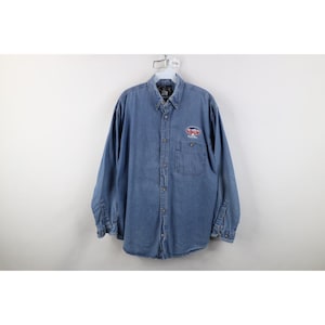 Pode incluir: Camisa de ganga de manga comprida em lavagem azul média. Possui gola abotoada, bolso no peito e um pequeno logotipo bordado no peito esquerdo. As mangas têm punhos abotoados.