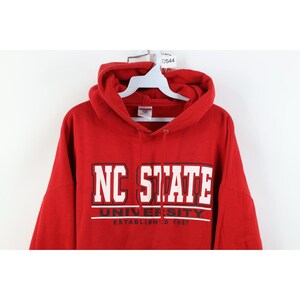 Peut inclure: Un sweat-shirt &agrave; capuche rouge avec le texte "NC STATE UNIVERSITY ESTABLISHED 1887" en lettres blanches. Le sweat-shirt a une capuche &agrave; cordon et une poche kangourou.