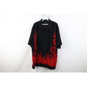 Pode incluir: Camisa preta de manga curta com botões e gola clássica. A camisa apresenta um design vibrante de chamas vermelhas nas laterais. Está pendurada em um cabide branco contra um fundo branco.