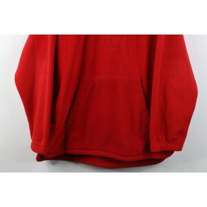 May include: A solid red fleece pullover with a kangaroo pocket and a small square patch near the neckline. The long sleeves have ribbed cuffs. The garment is made of a soft, warm material.