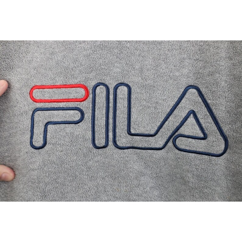 May include: Close-up of a gray fleece fabric with the FILA logo embroidered in navy blue and red. The logo is large and centered, with the brand name in a bold, outlined font. The fabric has a soft, textured appearance.