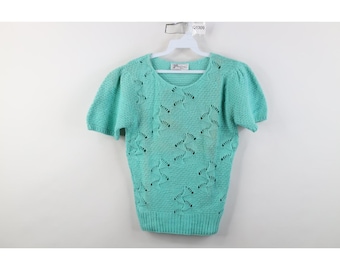 Puede incluir: Un suéter turquesa claro de manga corta con cuello redondo. El suéter presenta un diseño calado en forma de estrella en todo el cuerpo y mangas abullonadas. La prenda está colgada de una percha blanca.