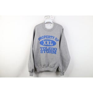 May include: A gray XXL sweatshirt with the text "PROPERTY OF" above an oval with "XXL" inside, and "TOLEDO STORM" below, all in blue. The sweatshirt is hanging on a white hanger against a white background.