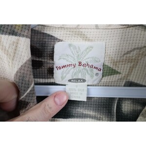 Op de afbeelding: Close-up van een Tommy Bahama zijden shirt label. Het label is wit met de merknaam en een palmboomafbeelding. Het shirt is maat S en gemaakt in China. Het shirt heeft een neutraal kleurenpalet.