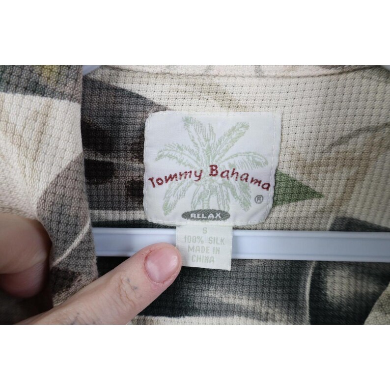 Op de afbeelding: Close-up van een Tommy Bahama zijden shirt label. Het label is wit met de merknaam en een palmboomafbeelding. Het shirt is maat S en gemaakt in China. Het shirt heeft een neutraal kleurenpalet.