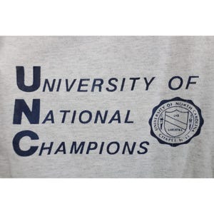 Op de afbeelding: Lichtgrijs t-shirt met de marineblauwe tekst "UNC University of National Champions". Aan de rechterkant van het shirt staat een marineblauw zegel met tekst en een schild.