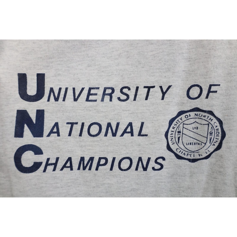 Op de afbeelding: Lichtgrijs t-shirt met de marineblauwe tekst "UNC University of National Champions". Aan de rechterkant van het shirt staat een marineblauw zegel met tekst en een schild.