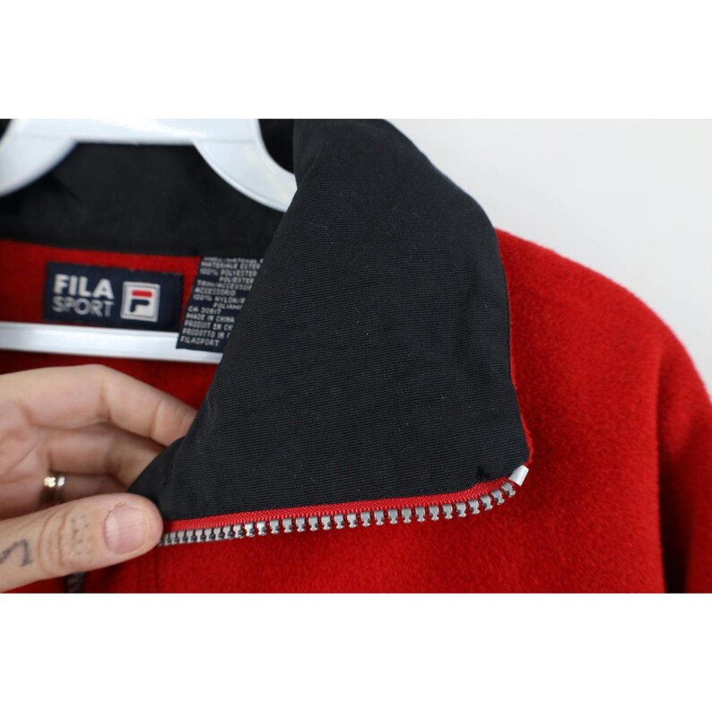May include: Red fleece jacket with a black collar and a silver zipper. The jacket has the "FILA SPORT" logo on the chest. The jacket is made of a soft, warm material.