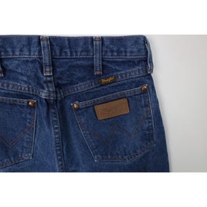 Puede incluir: Primer plano de la parte trasera de unos vaqueros azules. Los vaqueros tienen dos bolsillos traseros, remaches de color cobre y una etiqueta de cuero marr&oacute;n con el logotipo "Wrangler". Las costuras son de color beige en contraste.