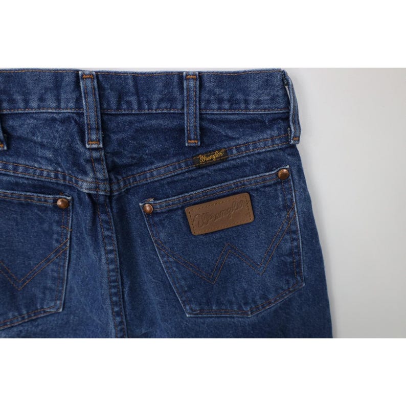 Puede incluir: Primer plano de la parte trasera de unos vaqueros azules. Los vaqueros tienen dos bolsillos traseros, remaches de color cobre y una etiqueta de cuero marr&oacute;n con el logotipo "Wrangler". Las costuras son de color beige en contraste.