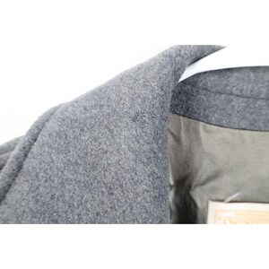 May include: Close-up of a gray wool coat collar. The coat has a textured surface and a light gray lining. The collar is folded over a white hanger.