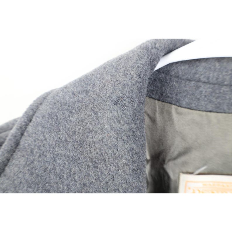 May include: Close-up of a gray wool coat collar. The coat has a textured surface and a light gray lining. The collar is folded over a white hanger.