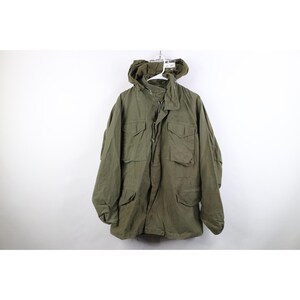 May include: An olive green field jacket featuring a hood, a front zipper closure, and four large button-flap pockets on the chest and waist. The jacket has long sleeves with adjustable cuffs and a structured collar, providing a utilitarian design.