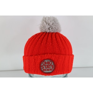 May include: A bright red ribbed knit beanie featuring a gray pom-pom on top. The front of the hat displays a circular patch with the text OHIO STATE BUCKEYES embroidered in red lettering against a gray background, centered on the folded cuff.
