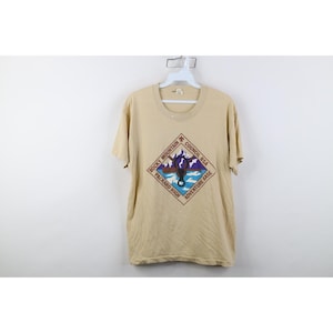May include: Tan short-sleeved t-shirt with a crew neck. The front features a diamond-shaped graphic with text that reads "Rocky Mountain Packard High Council BSA Adventure Base" surrounding a mountain and water scene.