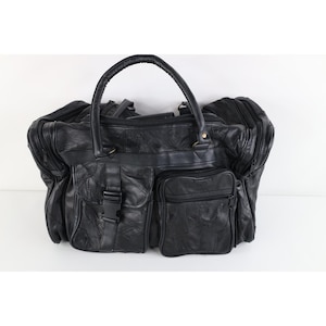 May include: A black leather duffel bag featuring a patchwork design, a top handle, and multiple exterior compartments. The front includes a pocket with a plastic buckle closure and a separate zippered pocket, with additional side zippered sections.