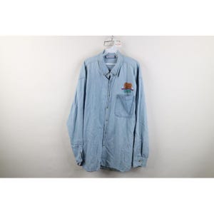 May include: A light blue long-sleeve denim button-down shirt featuring a single chest pocket. An embroidered graphic on the pocket displays a brown bear illustration above the text "GRIZZLY" in teal letters with a red banner accent.