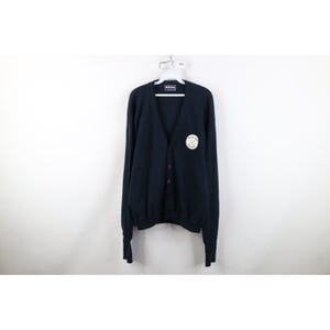 May include: A navy blue cardigan sweater with a V-neck and button closure. The sweater has a white and gold emblem on the left chest. The sweater is hanging on a white hanger against a white background.