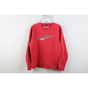 Könnte beinhalten: Ein rotes Guess Jeans Sweatshirt mit langen Ärmeln. Der Markenname ist in Schreibschrift in Weiß über der Brust geschrieben. Das Sweatshirt hängt an einem weißen Kleiderbügel.