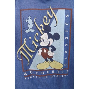 Op de afbeelding: Een blauw sweatshirt met een afbeelding van Mickey Mouse. Het ontwerp bevat de tekst "Mickey", "Original", "Classics", "Authentic" en "Finest in Quality" in een rechthoekig kader met een lichtblauwe diagonale streep als achtergrond.