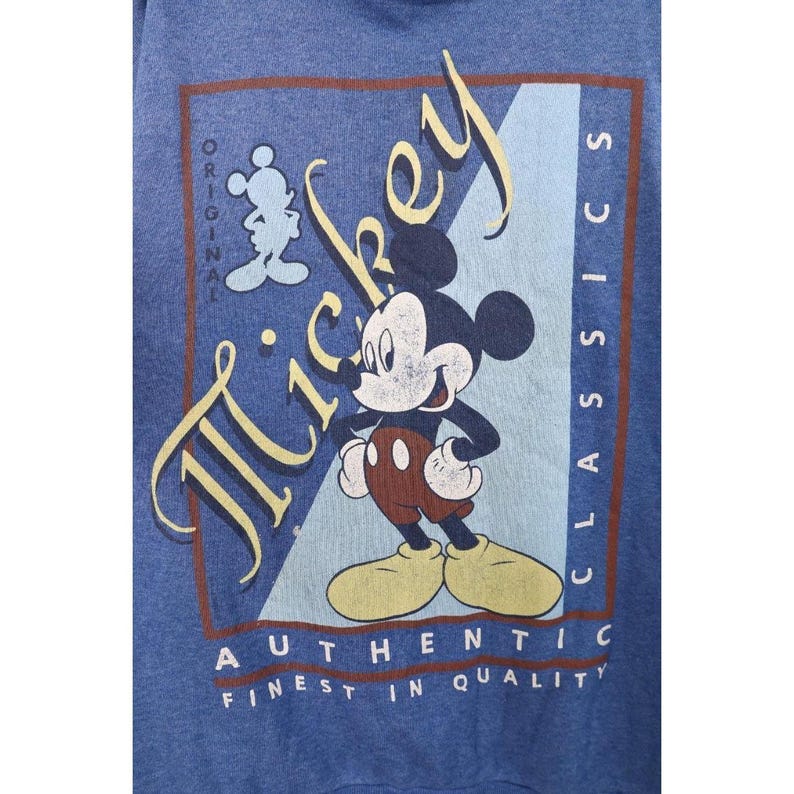 Op de afbeelding: Een blauw sweatshirt met een afbeelding van Mickey Mouse. Het ontwerp bevat de tekst "Mickey", "Original", "Classics", "Authentic" en "Finest in Quality" in een rechthoekig kader met een lichtblauwe diagonale streep als achtergrond.