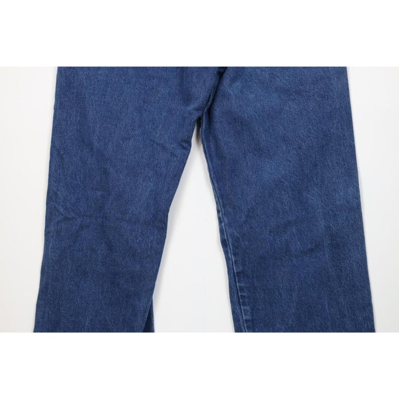 May include: Close-up of blue denim jeans. The jeans have a classic five-pocket design and a straight leg cut. The denim has a medium wash with some fading and wear.