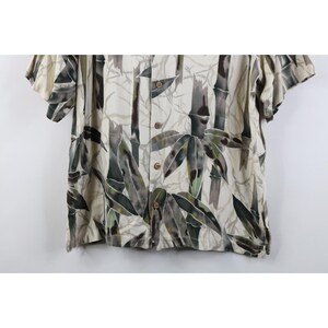 Op de afbeelding: Korte mouwen overhemd met bamboe print. Het shirt heeft een cr&egrave;mekleurige achtergrond met een patroon van groene, bruine en grijze bamboestengels en bladeren. Het heeft een klassieke kraag en houten knopen.