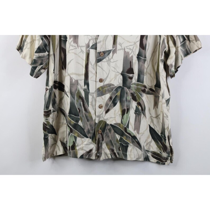 Op de afbeelding: Korte mouwen overhemd met bamboe print. Het shirt heeft een cr&egrave;mekleurige achtergrond met een patroon van groene, bruine en grijze bamboestengels en bladeren. Het heeft een klassieke kraag en houten knopen.