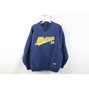 Op de afbeelding: Een marineblauw sweatshirt met gele "Michigan"-letters en een "M"-logo. Het sweatshirt heeft een V-hals en een klein label aan de linkerkant. Het hangt aan een witte hanger.