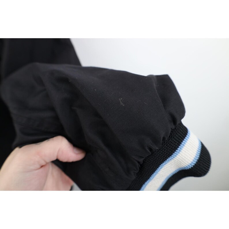 May include: Black jacket sleeve with a ribbed cuff. The cuff features a black base with white and light blue stripes. The jacket appears to be made of a smooth, possibly nylon, material.