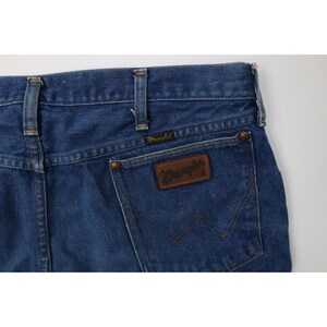 Op de afbeelding: Close-up van de achterkant van een blauwe Wrangler jeans van denim. De jeans heeft een bruine leren patch met het Wrangler logo en een klassiek vijf-zakken ontwerp. De jeans heeft een medium wassing.