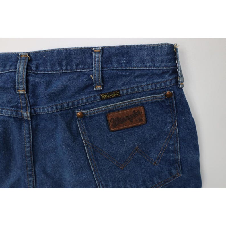 Op de afbeelding: Close-up van de achterkant van een blauwe Wrangler jeans van denim. De jeans heeft een bruine leren patch met het Wrangler logo en een klassiek vijf-zakken ontwerp. De jeans heeft een medium wassing.
