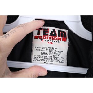 May include: A black Team Edition Apparel XXL jacket label. The label is white with red and black text, listing the shell and lining materials. The jacket is made in Sri Lanka.